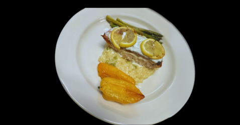 Grilled fish with lemon slices, asparagus, and yellow bell pepper on creamy risotto, served on a white plate.