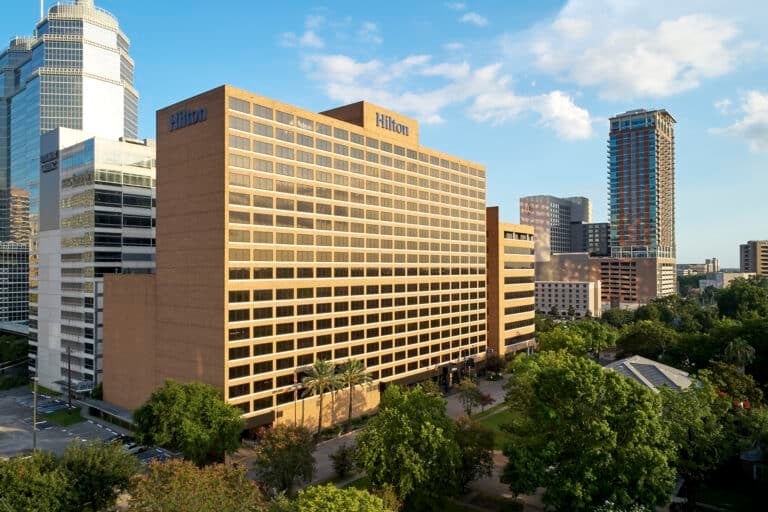 A large Hilton hotel building with a tan facade is surrounded by trees and other modern high-rise buildings under a partly cloudy sky in an urban setting.