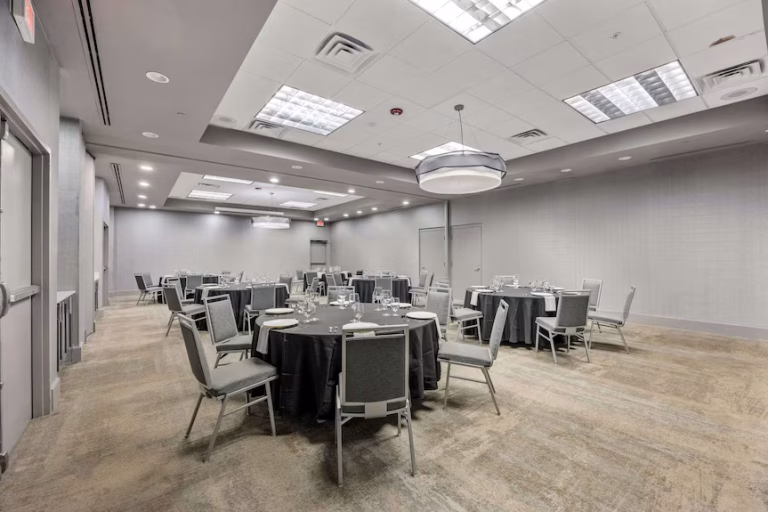 Spacious conference room with round tables covered in dark tablecloths, set with plates and glasses. Gray chairs surround each table. The room has neutral walls, a patterned carpet, and bright ceiling lights.