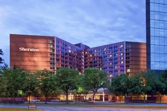 Exterior view of the Sheraton hotel at dusk, with warm lights glowing from the windows, surrounded by trees and a glass building on the right. The Sheraton sign is lit up on the left side of the hotel.