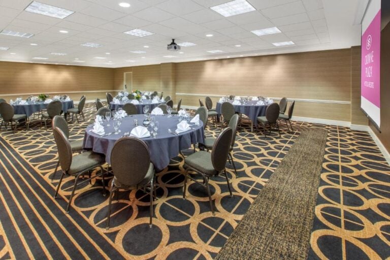 A modern banquet room with round tables covered in gray tablecloths, set with glassware and white napkins. Patterned carpet, beige walls, and a projection screen are visible in the brightly lit space.