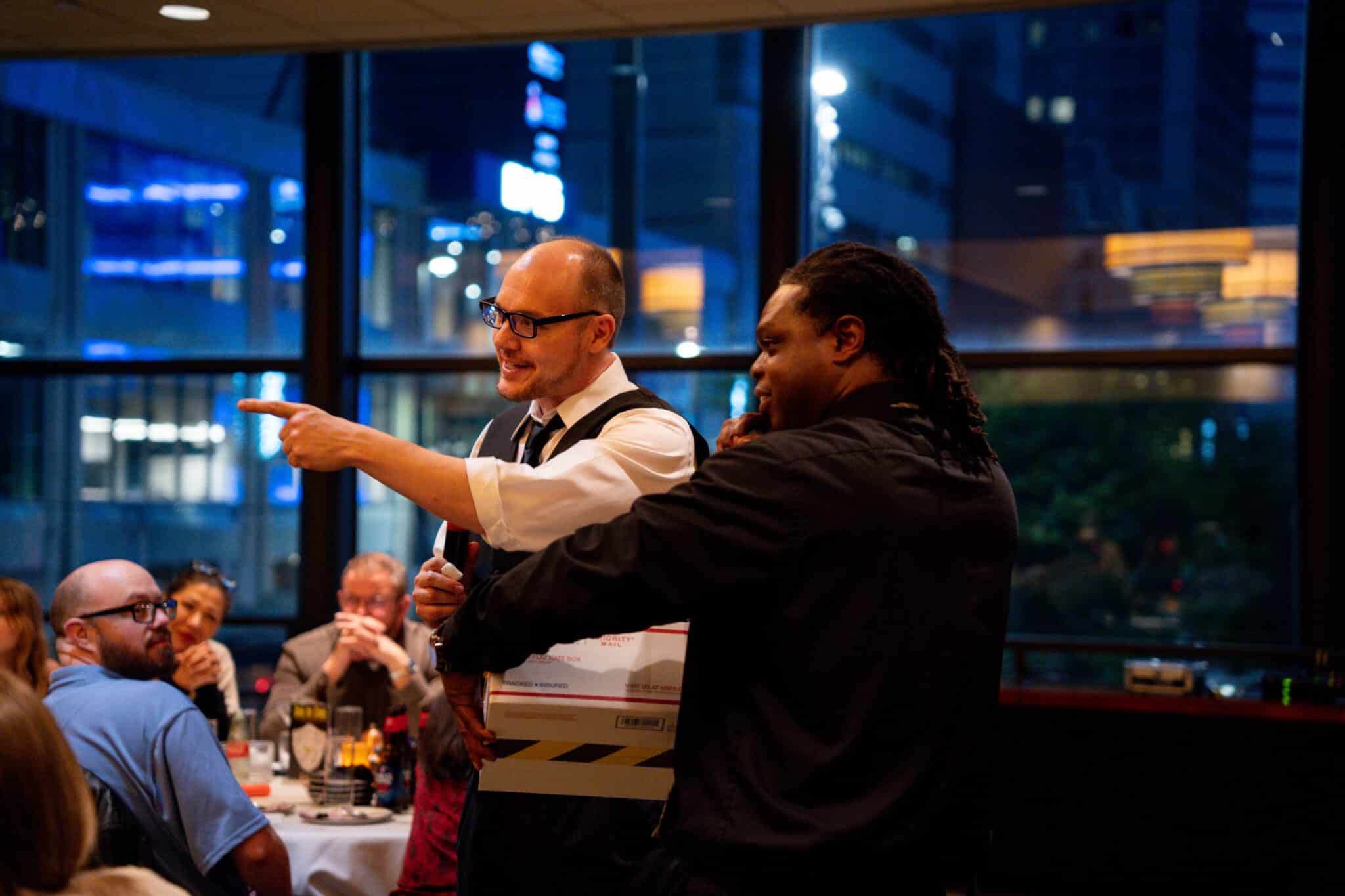 Two guests standing and pointing dramatically during an interactive murder mystery dinner show, with a nighttime city skyline visible through the windows behind them
