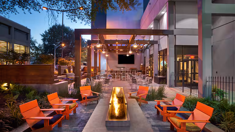 Modern outdoor patio with orange cushioned chairs around a long fire pit, tables and chairs under a pergola, and string lights above. The area is adjacent to a contemporary building at dusk.