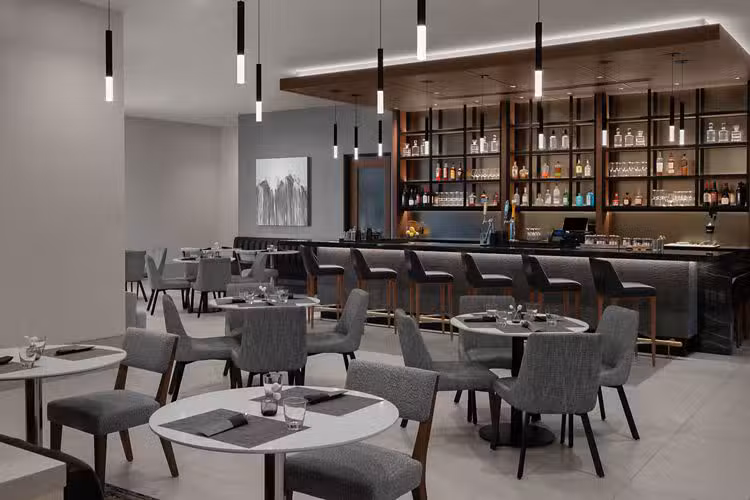 Modern restaurant interior with gray chairs and round tables, place settings ready. A sleek bar with shelves of bottles and glasses is in the background. Contemporary pendant lights hang from the ceiling.