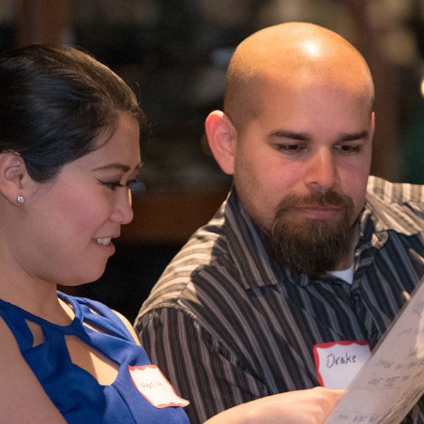 A man and woman are sitting together, looking at a piece of paper. The man is bald with a beard, wearing a dark shirt. The woman has long dark hair, wearing a blue sleeveless top. Both have name tags. Another person is partially visible on the left.