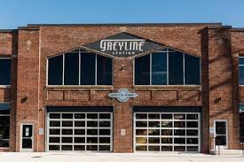 A brick building in Lexington, KY, showcases large garage-style doors and a Greyline Station sign with Crafts & Vines below. Large windows grace the upper part against a clear blue sky, making it a must-see spot when exploring things to do in Lexington, KY.