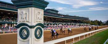 A horse race in progress at a Lexington racetrack captivates spectators in the stands. The foreground showcases a decorative pillar, while the background highlights a large building against a clear blue sky—one of the many exhilarating things to do in Lexington, KY.