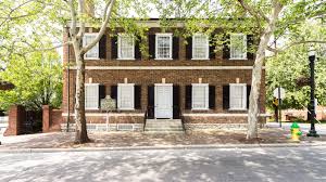 A charming two-story brick building with white shutters and doors is nestled among green trees in Lexington, KY. In the foreground, a sidewalk and street accommodate leisurely strolls past a fire hydrant and streetlamp, offering one of many pleasant things to do in Lexington.