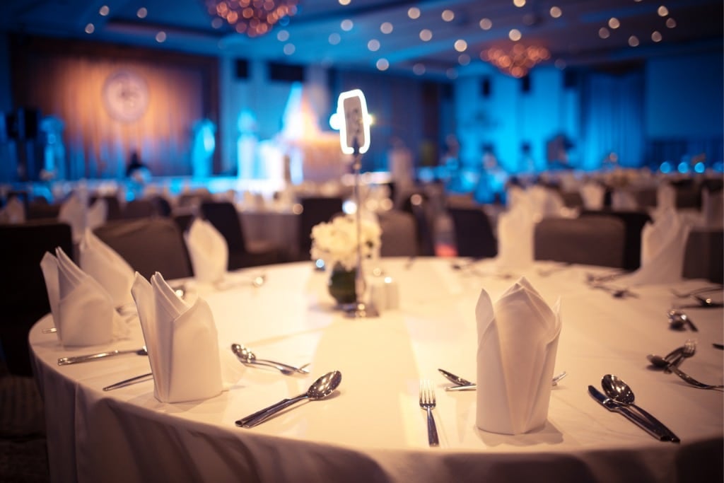 A round banquet table is elegantly set in a dimly lit event hall at Hilton Lexington Downtown. It features neatly folded white napkins, silver cutlery, and a small centerpiece. The background is a blur of blue lighting and chandeliers, creating a formal atmosphere perfect for exclusive events.