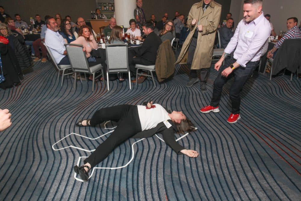 A staged crime scene at The Dinner Detective murder mystery event, with an actor playing the victim while guests and detectives investigate.