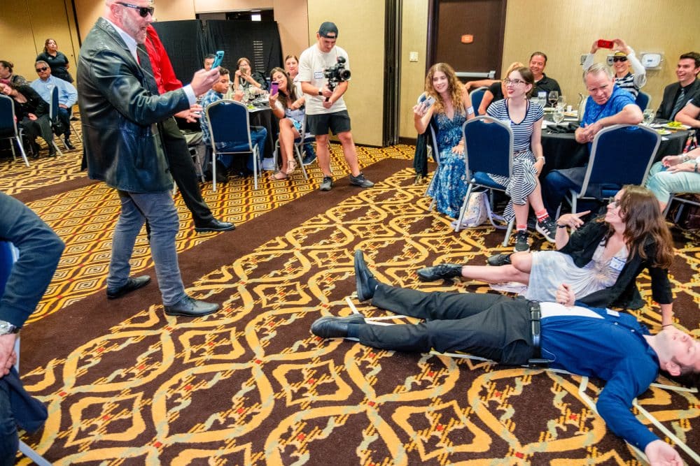 A lively scene at an indoor event. A man in a leather jacket is holding a microphone as a person lies flat on the patterned carpet. Attendees in casual and semi-formal attire watch and laugh, while some take photos. Blue chairs and tables surround the area.