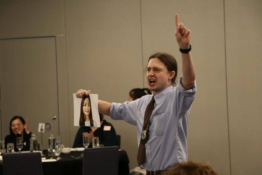 An actor playing a detective at The Dinner Detective murder mystery event in Lexington, KY, holding up a suspect's photo while engaging the audience.