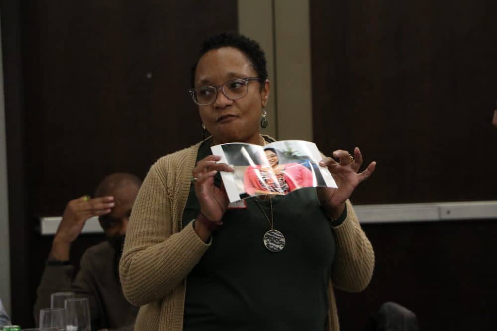A person wearing glasses and a beige cardigan holds up a folded photo while speaking. They stand in a room with another individual, possibly discussing plans for the Lexington murder mystery dinner show as an intriguing evening idea.