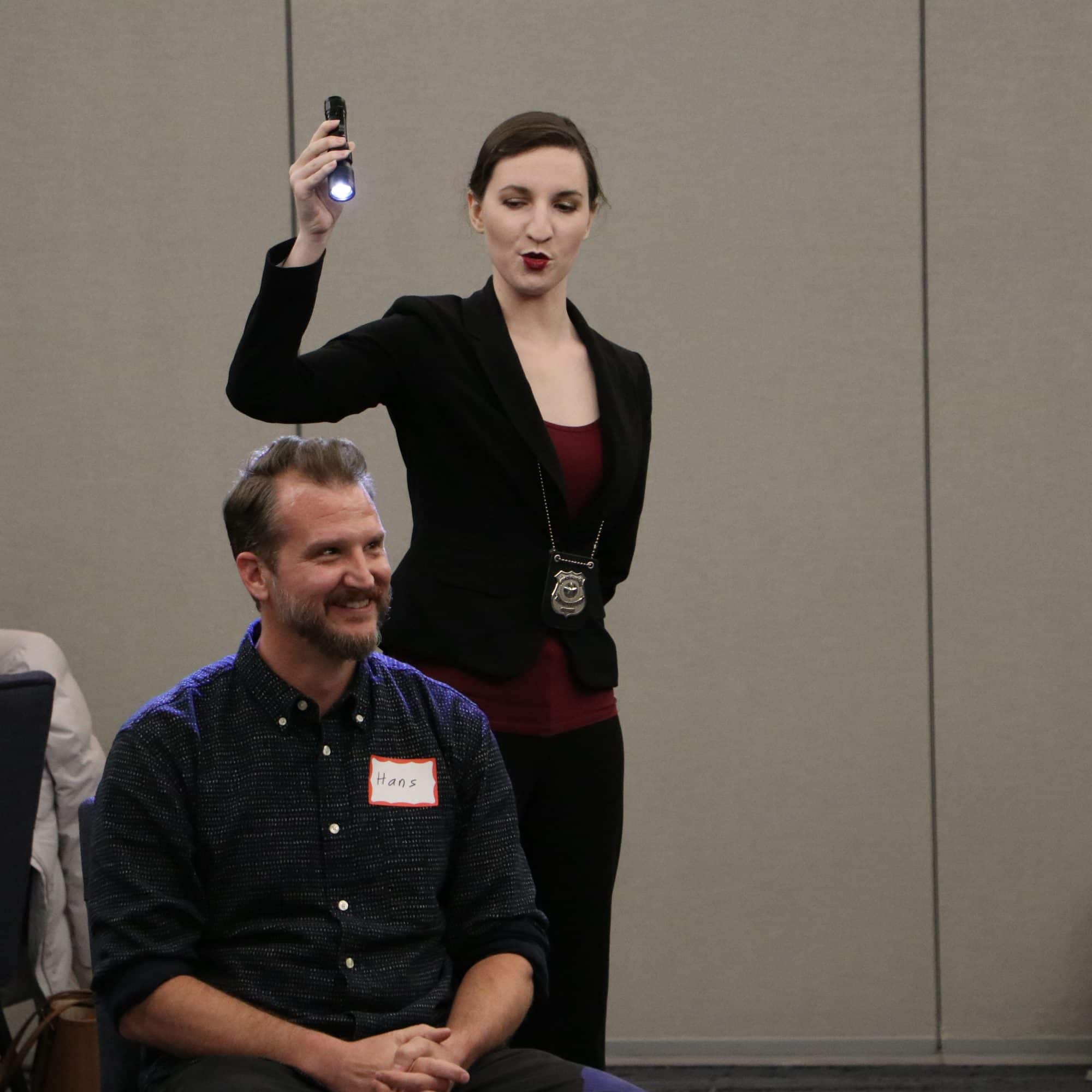 A woman in a black suit stands holding a flashlight during a presentation on romantic things to do in Lexington, smiling confidently. A man with a beard sits in front of her, also smiling. Other people are seated in the background, perhaps planning their own adventures.