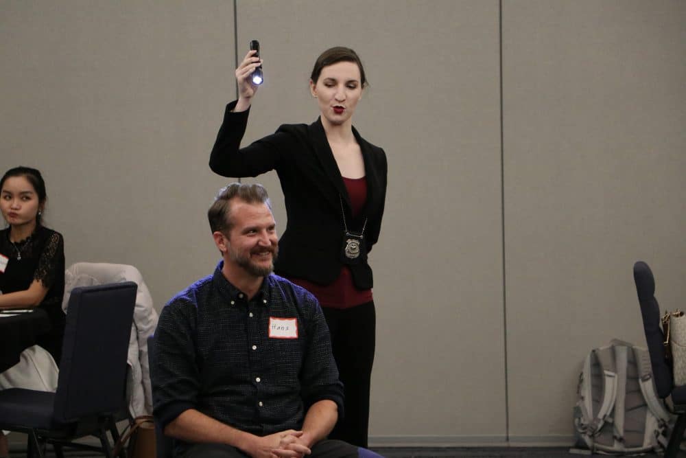 A detective character at The Dinner Detective murder mystery event in Lexington, KY, holding up a flashlight while engaging with a participant.