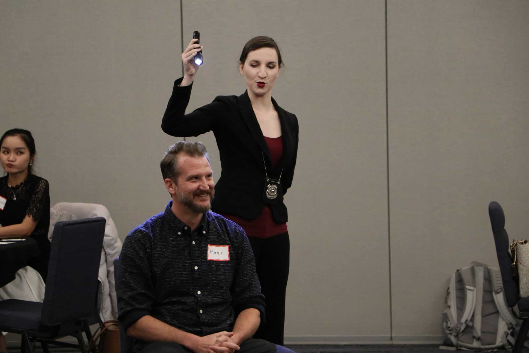 A detective character at The Dinner Detective murder mystery event in Lexington, KY, holding up a flashlight while engaging with a participant.