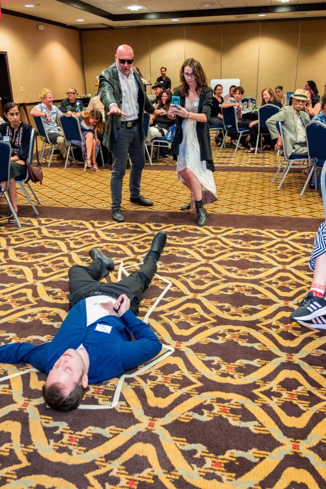 A lively event scene in a conference room with people watching. A person in a costume dramatically points at another lying on the floor within a chalk outline. A woman in a costume takes a photo. People are seated in chairs observing the spectacle.