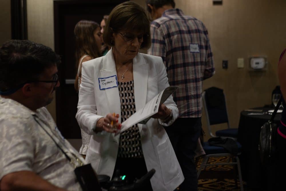 A woman in a white blazer, wearing glasses and a name tag, examines a paper at a social gathering. A man in a patterned shirt sits nearby, and others are conversing in the background. The setting appears to be an indoor event.
