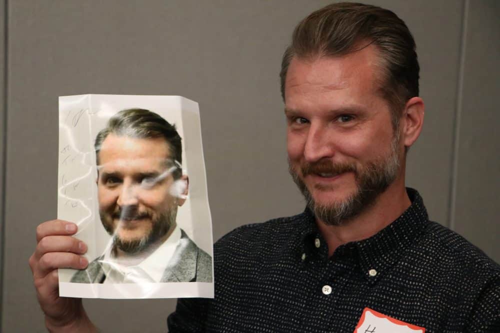 A man with a beard is smiling and holding up a crumpled piece of paper with a photo of himself printed on it. He is wearing a dark, dotted shirt and standing against a plain background.