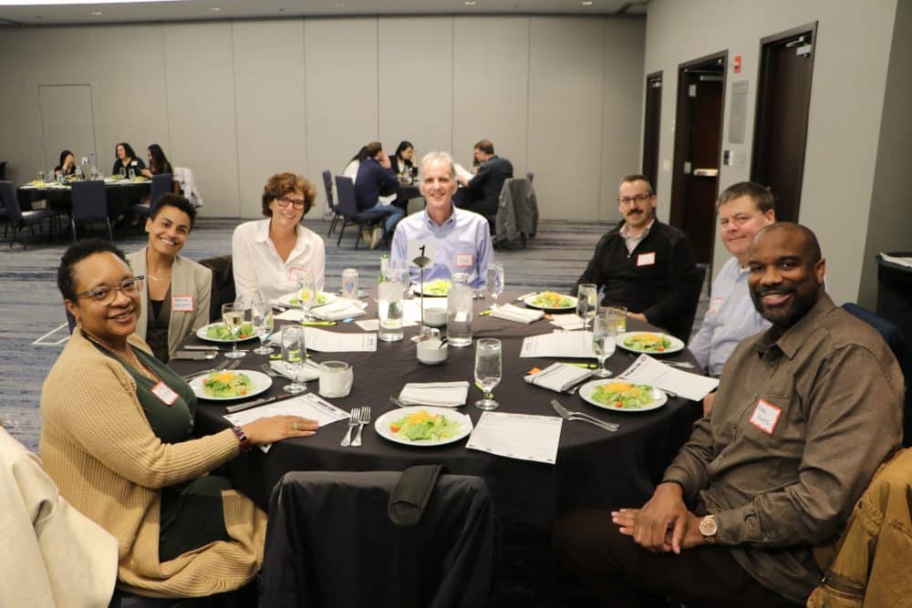 A group of seven people sit around a round table in a conference room, smiling at the camera. They have plates of salad in front of them. The room has several tables with people in the background.