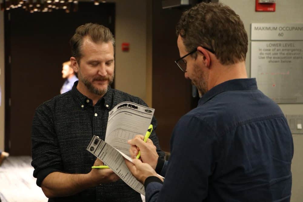Two men are engaged in discussion indoors. One is holding a clipboard and pen, while the other looks on. They appear to be examining documents. The setting seems professional, with signs and a dimly lit background.