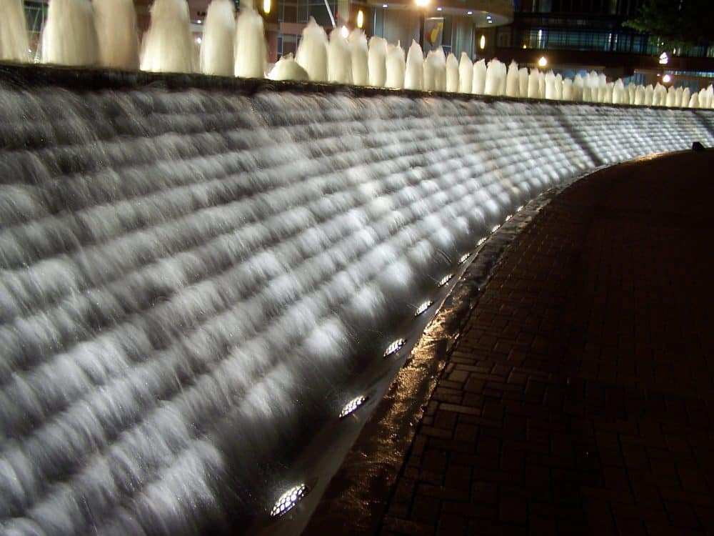 A curved fountain with cascading water, illuminated by lights at night. The water flows smoothly over textured stone, creating a glowing effect. The surrounding area is paved with bricks, and lights are visible in the background.