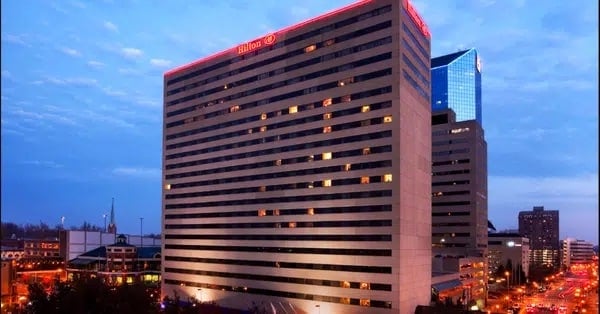 A tall Hilton hotel building at dusk, with illuminated windows and a glowing red sign on top. City streets with lights and other buildings are visible in the background under a blue sky.