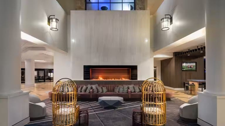 A modern hotel lobby with a large fireplace, brown sectional sofa, patterned cushions, and two yellow birdcage chairs. Warm lighting and neutral tones create a cozy, inviting atmosphere.