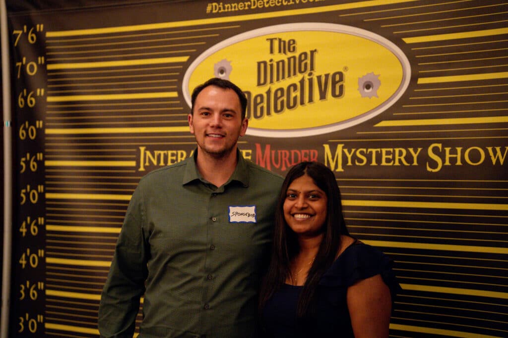 Two smiling guests posing in front of The Dinner Detective photo backdrop at a live interactive murder mystery event in Lexington