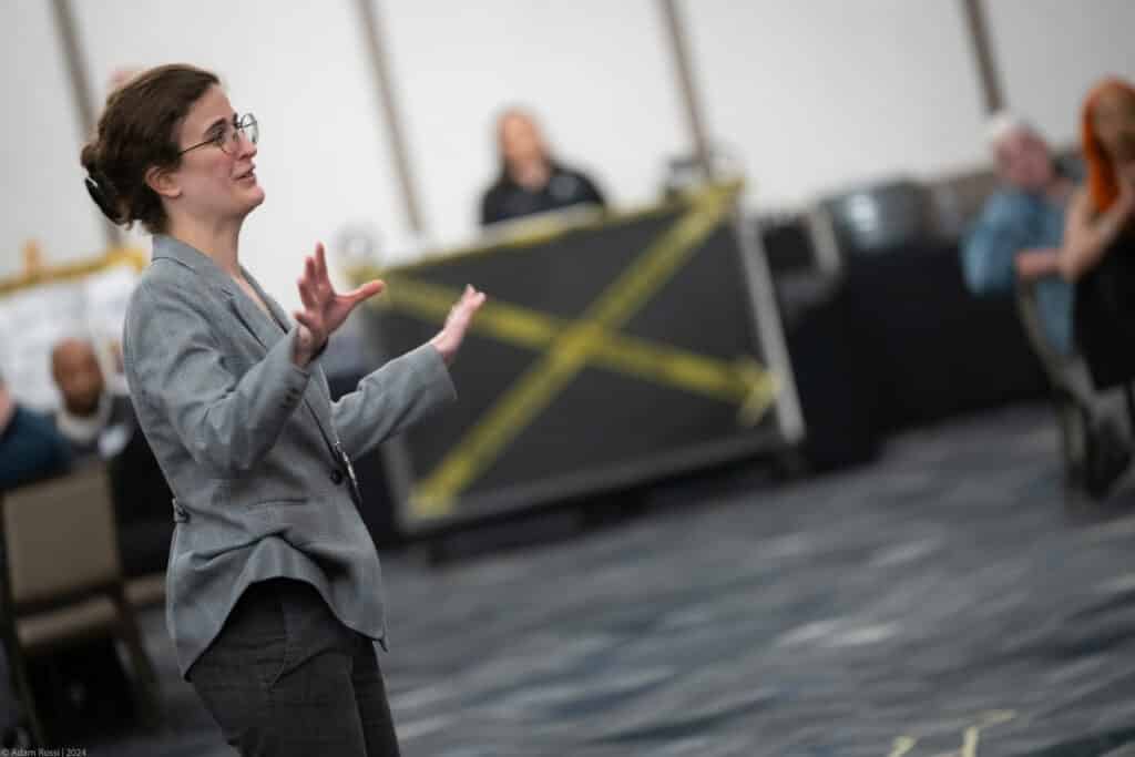 Female actor in a gray blazer gestures dramatically during a live performance at The Dinner Detective show.