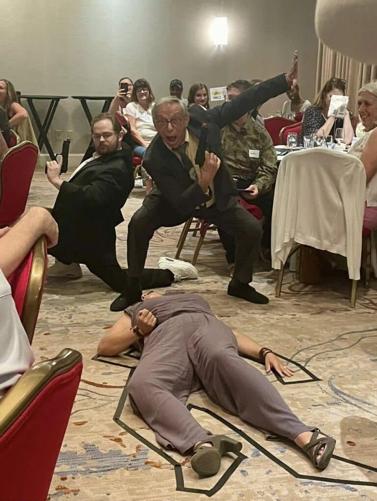 Actors performing a dramatic murder scene at The Dinner Detective with a victim on the floor inside a body outline while guests laugh and watch