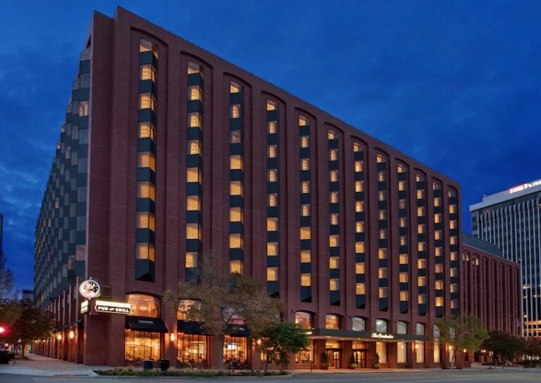 A large, modern brick hotel building with many lit windows at night, featuring a well-lit entrance and shops on the ground floor, set against a deep blue sky.