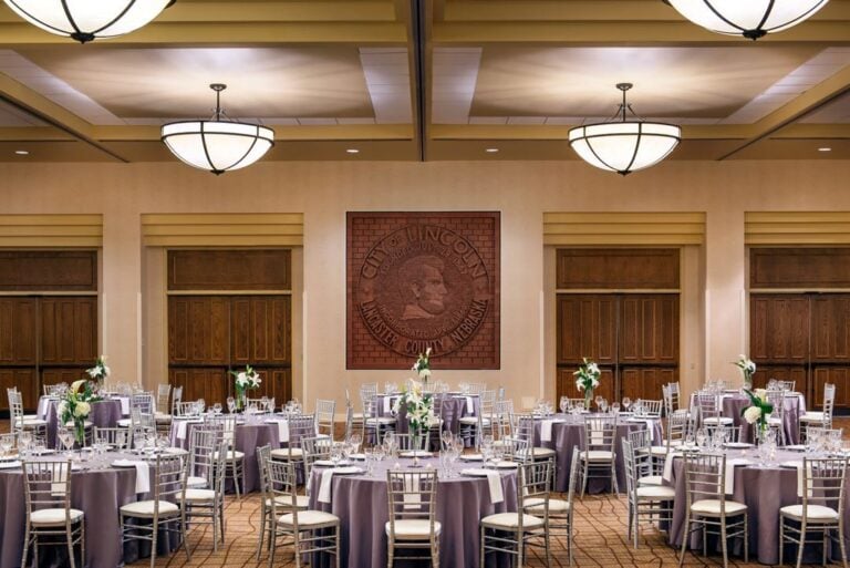 A banquet hall set for an event, featuring round tables with gray tablecloths, silver chairs, and floral centerpieces. Large decorative coin art with a profile is displayed on the far wall above wooden doors.