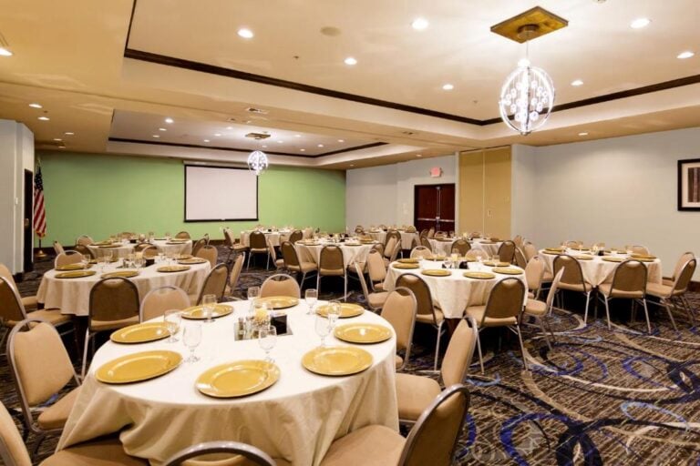 A banquet room with several round tables set with gold plates and glassware, surrounded by beige chairs. The room has a green accent wall, a large projection screen, and bright overhead lighting.