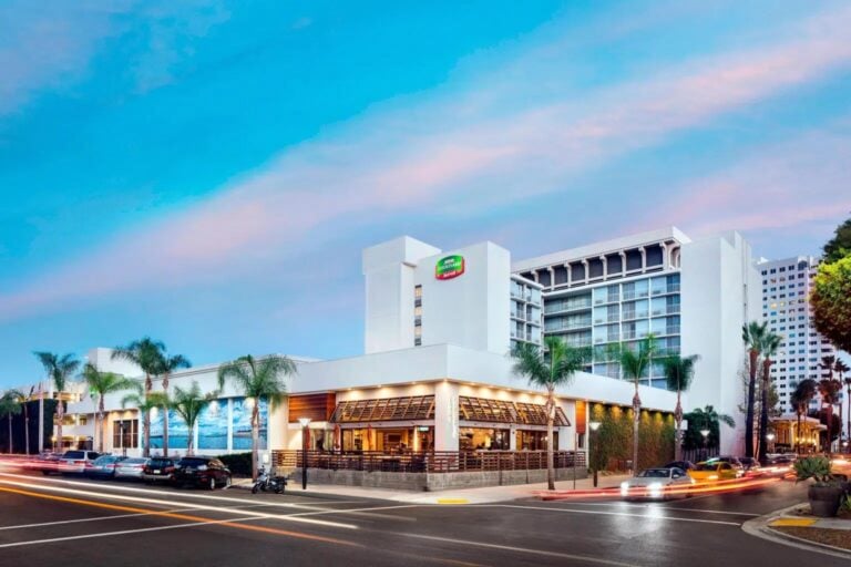 A modern hotel with white exterior, palm trees, and an outdoor patio is situated at a street corner, with blurred car lights passing by under a blue and pink sky at sunset.