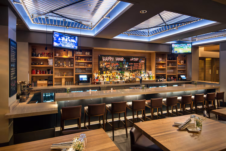 Modern bar with high ceilings, a long marble counter with barstools, shelves of liquor bottles, TV screens, and a chalkboard sign reading Happy Hour. Set tables with napkins and flowers in the foreground.
