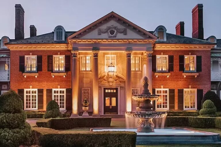 A grand, symmetrical red-brick mansion with tall columns, black shutters, and warm lighting stands behind a large, ornate fountain and manicured hedges at dusk.