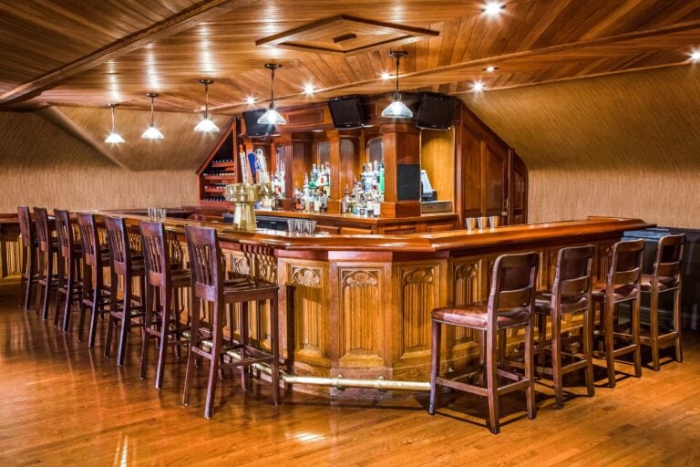 A wooden bar with high chairs lined up around it, shelves of bottles behind the counter, pendant lights overhead, and a polished wood floor and ceiling creating a warm, inviting atmosphere.