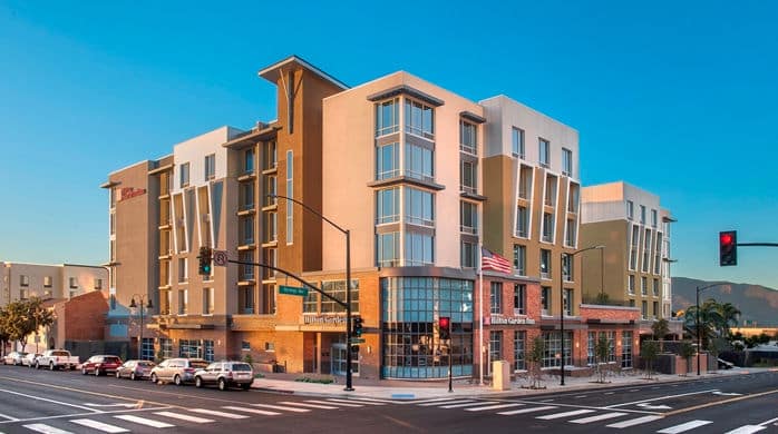 A modern multi-story hotel with large windows and a U.S. flag out front stands at a sunny street corner with parked cars and palm trees, under a clear blue sky.