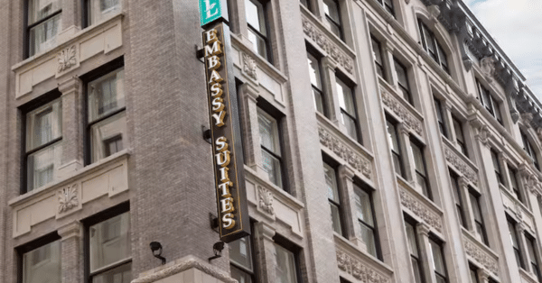 Exterior view of a brick building featuring a vertical Embassy Suites sign. The architecture includes ornate stonework around the tall windows. The sky is partially visible, suggesting daytime.
