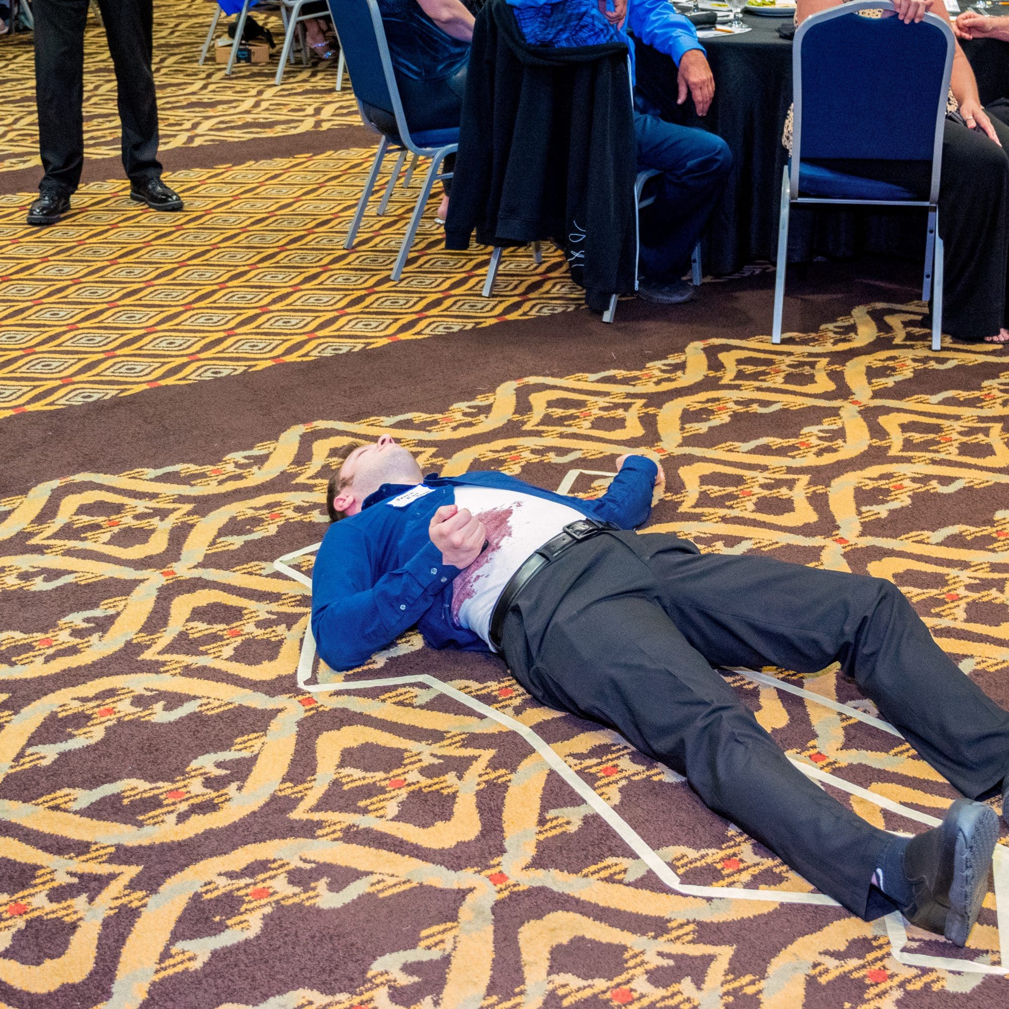 A man lies inside a taped outline on the floor, playfully acting as the “victim” during The Dinner Detective Murder Mystery Show in Louisville.