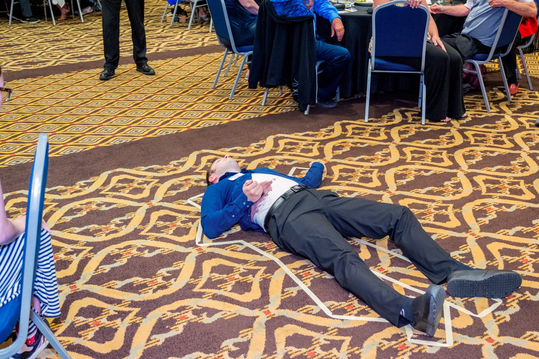 A man lies inside a taped outline on the floor, playfully acting as the “victim” during The Dinner Detective Murder Mystery Show in Louisville.