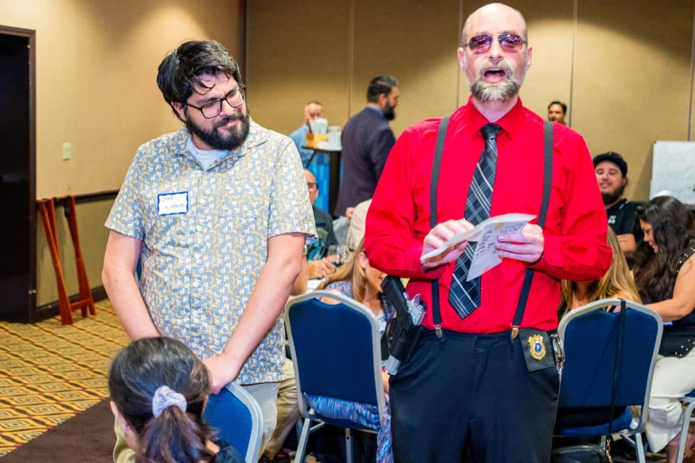 A performer in red suspenders and sunglasses speaks to the crowd while a guest reacts humorously during The Dinner Detective Show in Louisville.