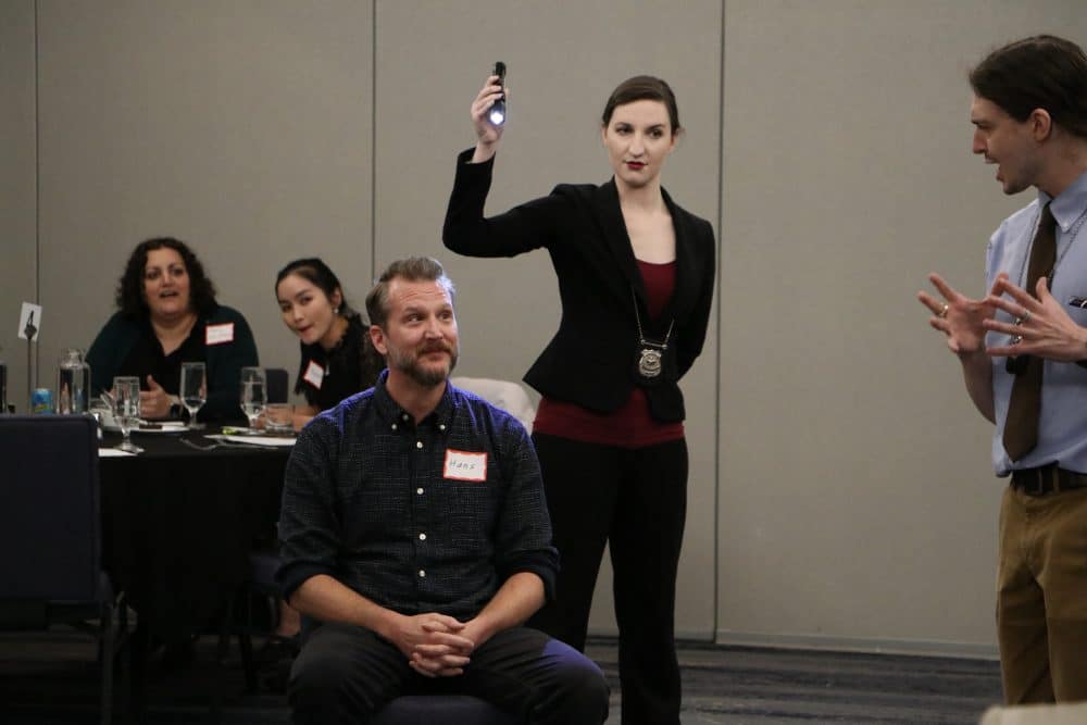 A woman in a detective costume shines a flashlight while interacting with seated dinner guests at a live mystery show.