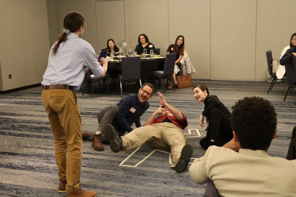 Guests laughing and posing next to a “crime scene” chalk outline during The Dinner Detective Murder Mystery Show in Louisville.