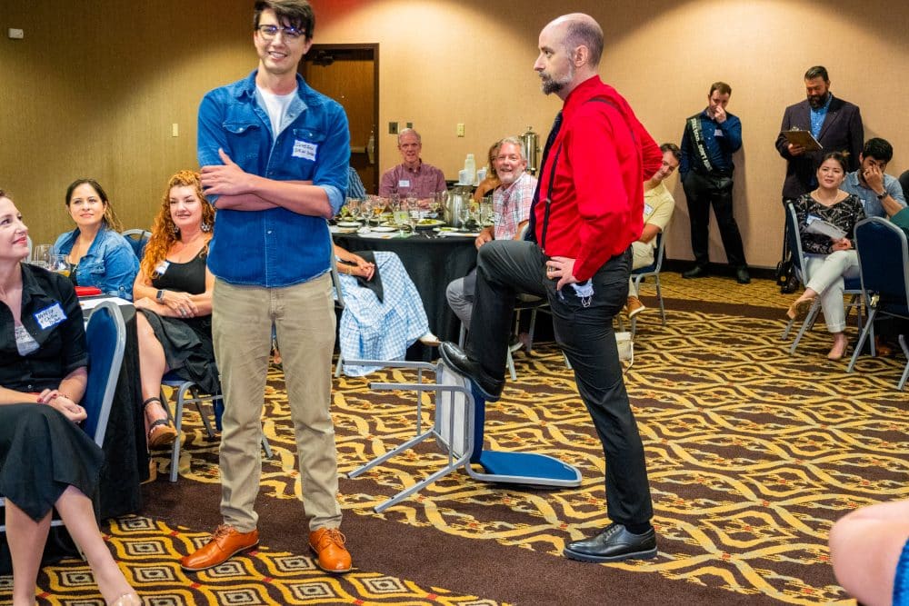 Two performers interact in character during a live Dinner Detective show as guests watch and laugh around them.