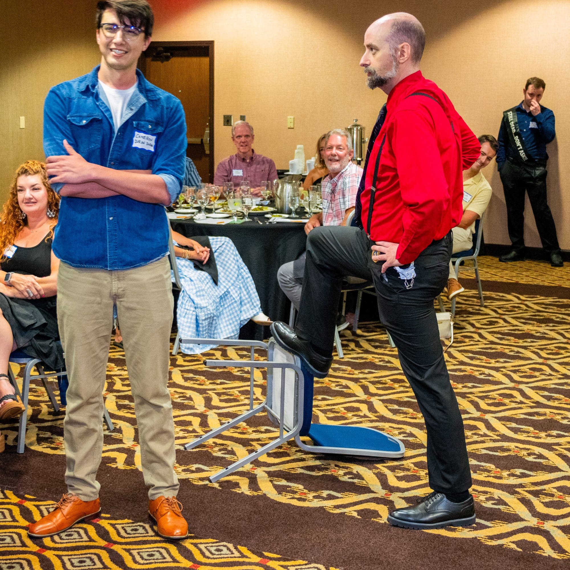 Two performers interact in character during a live Dinner Detective show as guests watch and laugh around them.