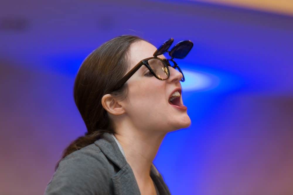 Female actor in costume, mid-performance, wearing quirky double sunglasses during a Dinner Detective comedy-mystery show.