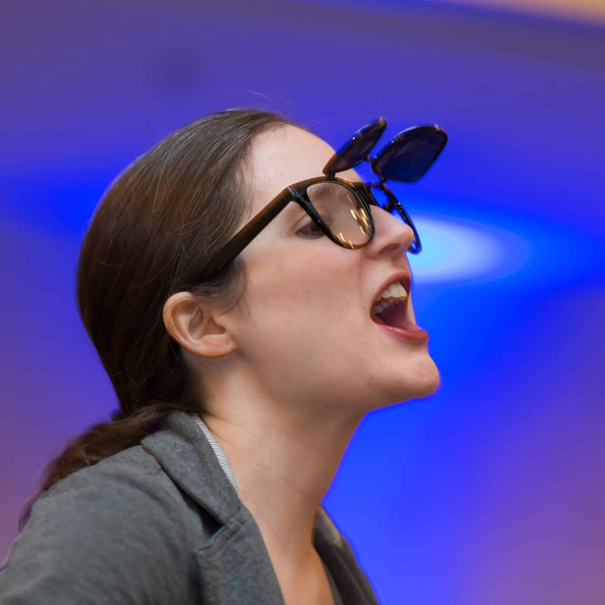 Female actor in costume, mid-performance, wearing quirky double sunglasses during a Dinner Detective comedy-mystery show.