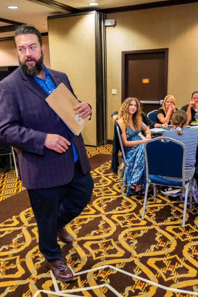 Performer with clipboard engaging audience at Dinner Detective event in a Louisville banquet room.